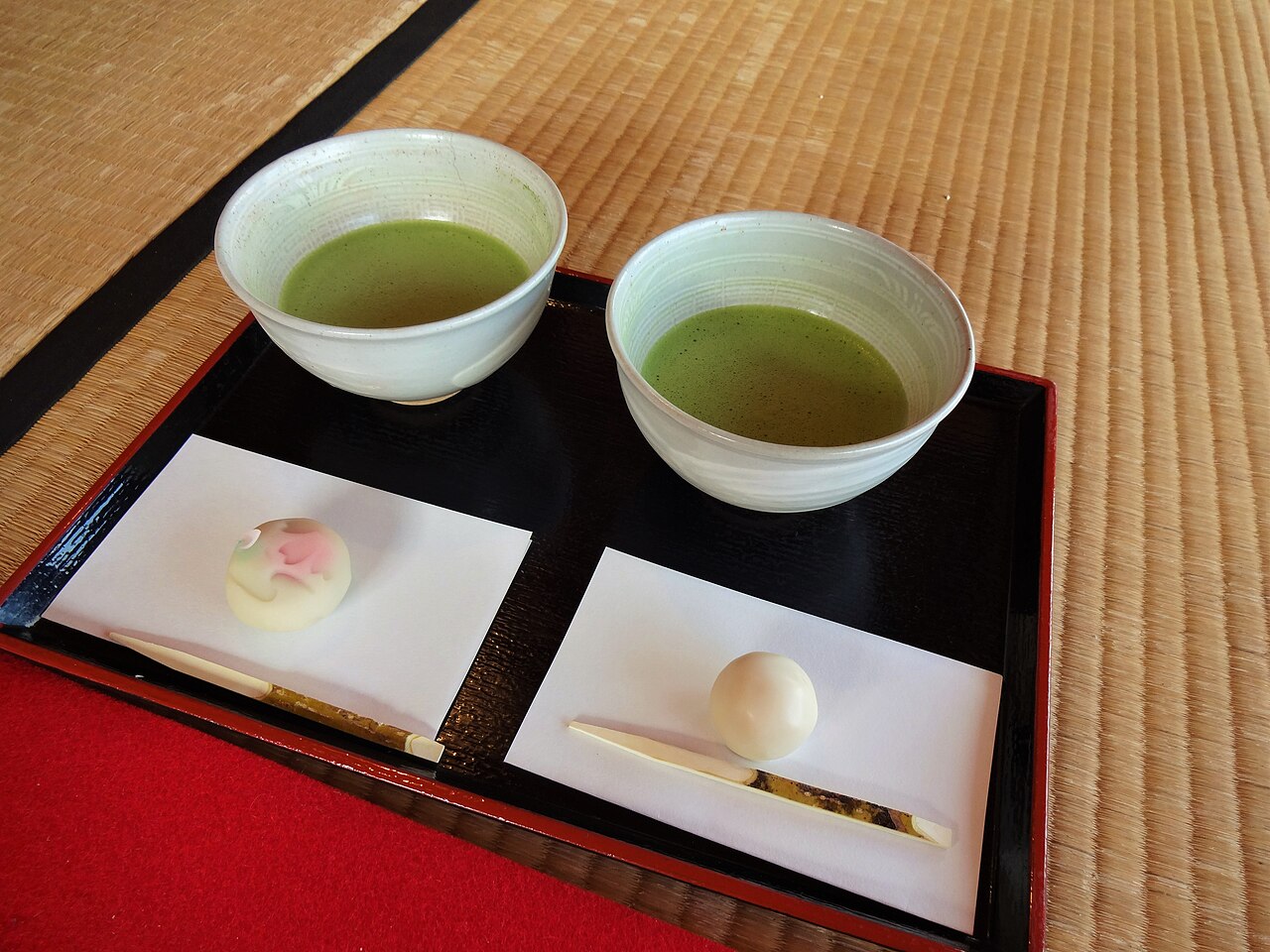 Two bowls of matcha on a black lacquer tray with wagashi sweets, set on a tatami mat