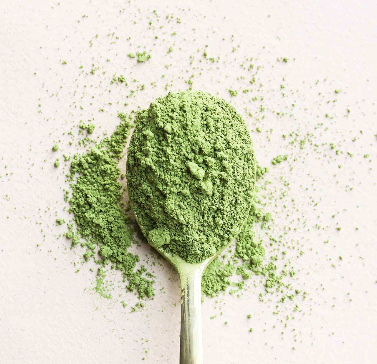 Spoon heaped with vivid green matcha powder scattered on a pale surface