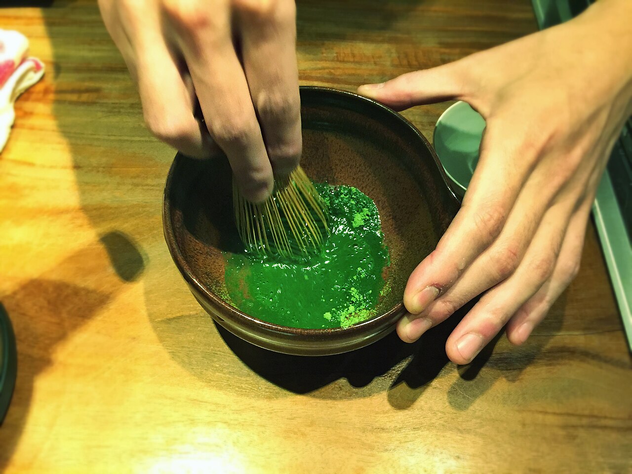 Hands whisking bright green matcha in a ceramic bowl with a bamboo whisk